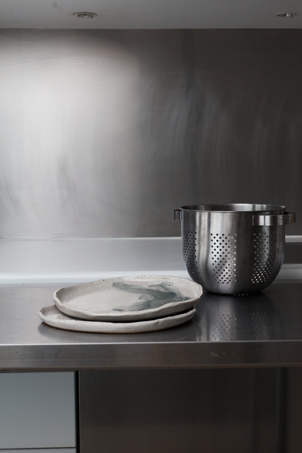 Metallic colander and plates on a stainless steel countertop with a metallic backsplash.