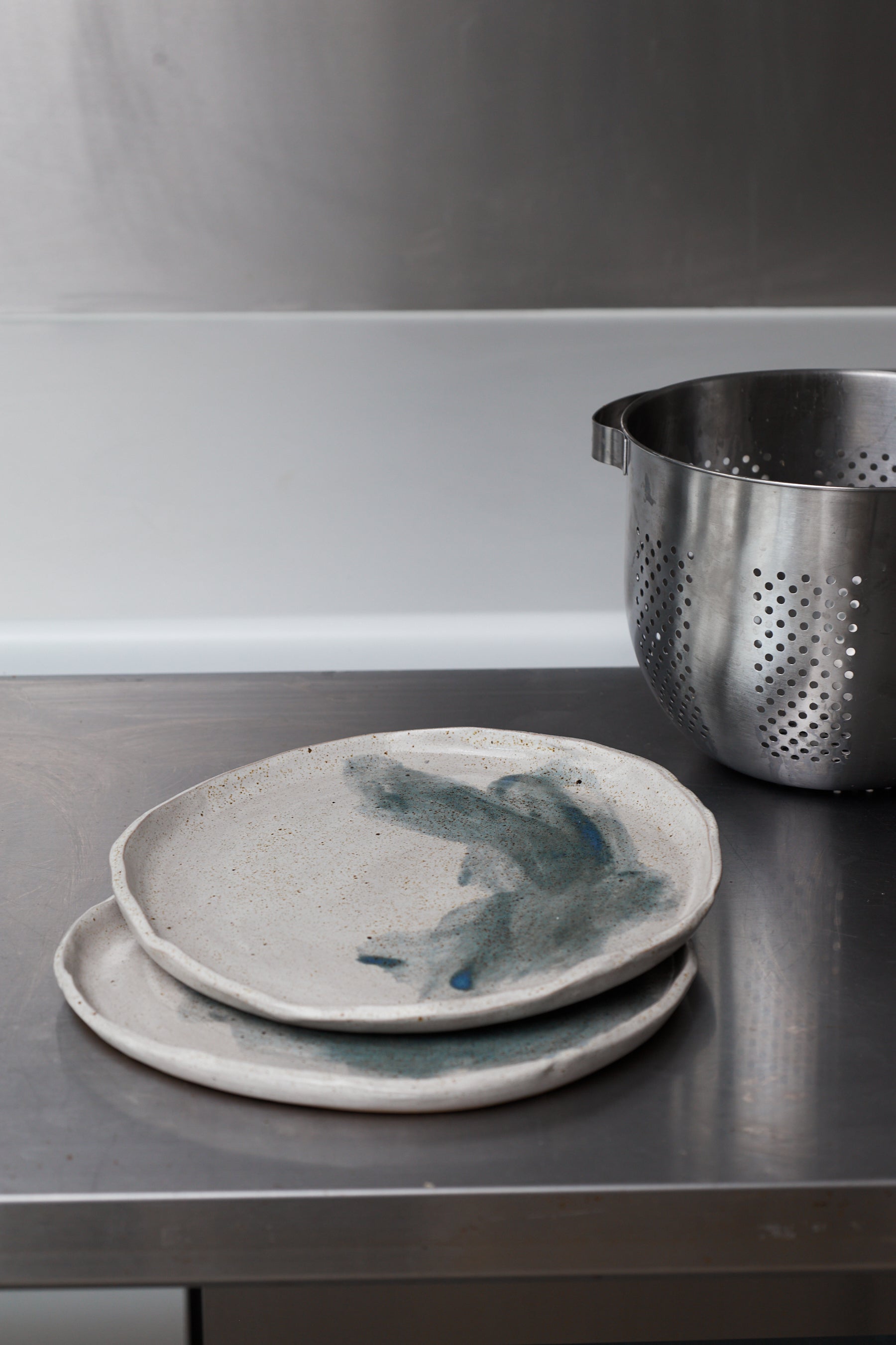 Two ceramic plates with blue abstract designs on a dark surface, with a metal colander in the background.