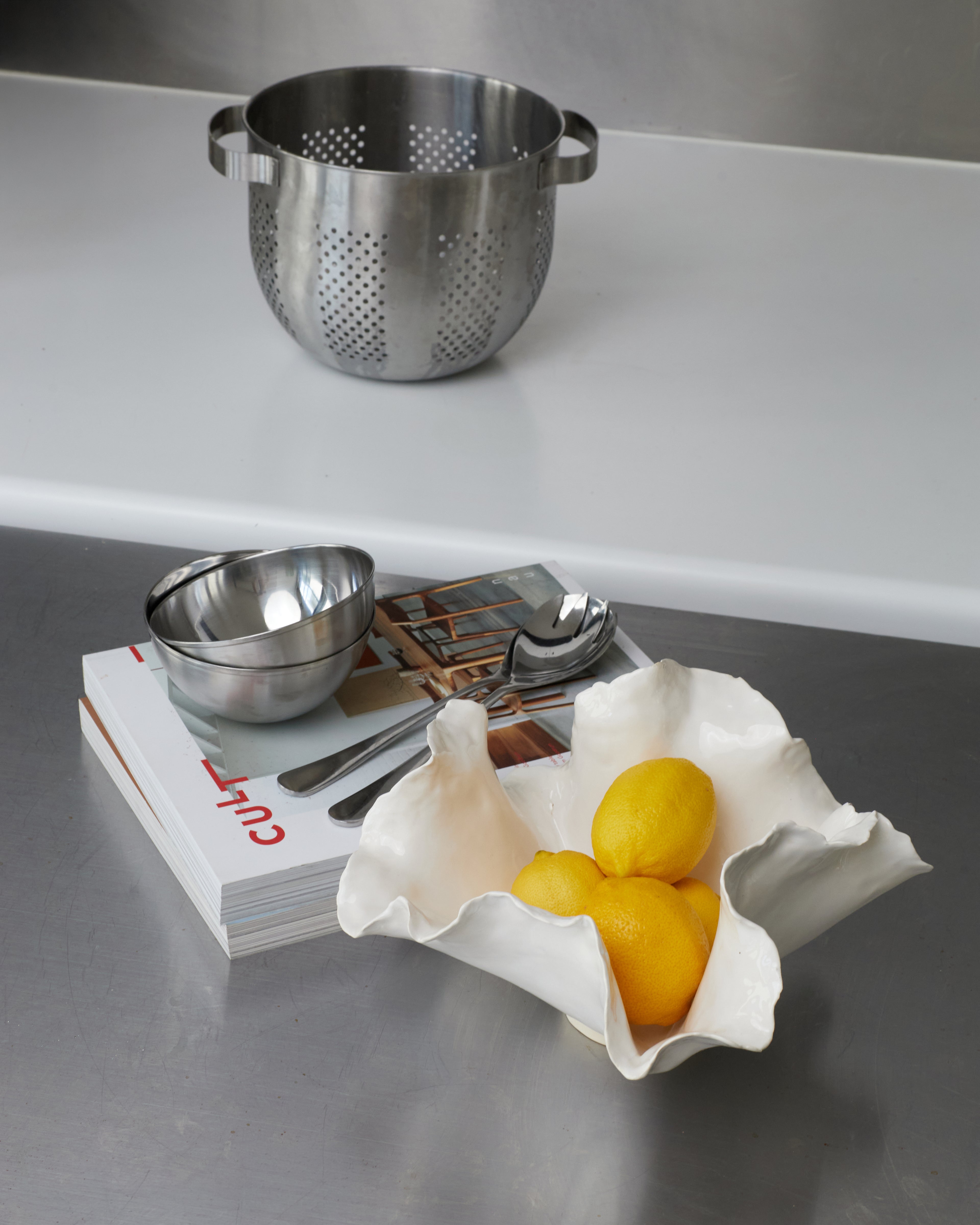 Metal colander and bowls on a magazine with lemons in a white dish on a metallic surface