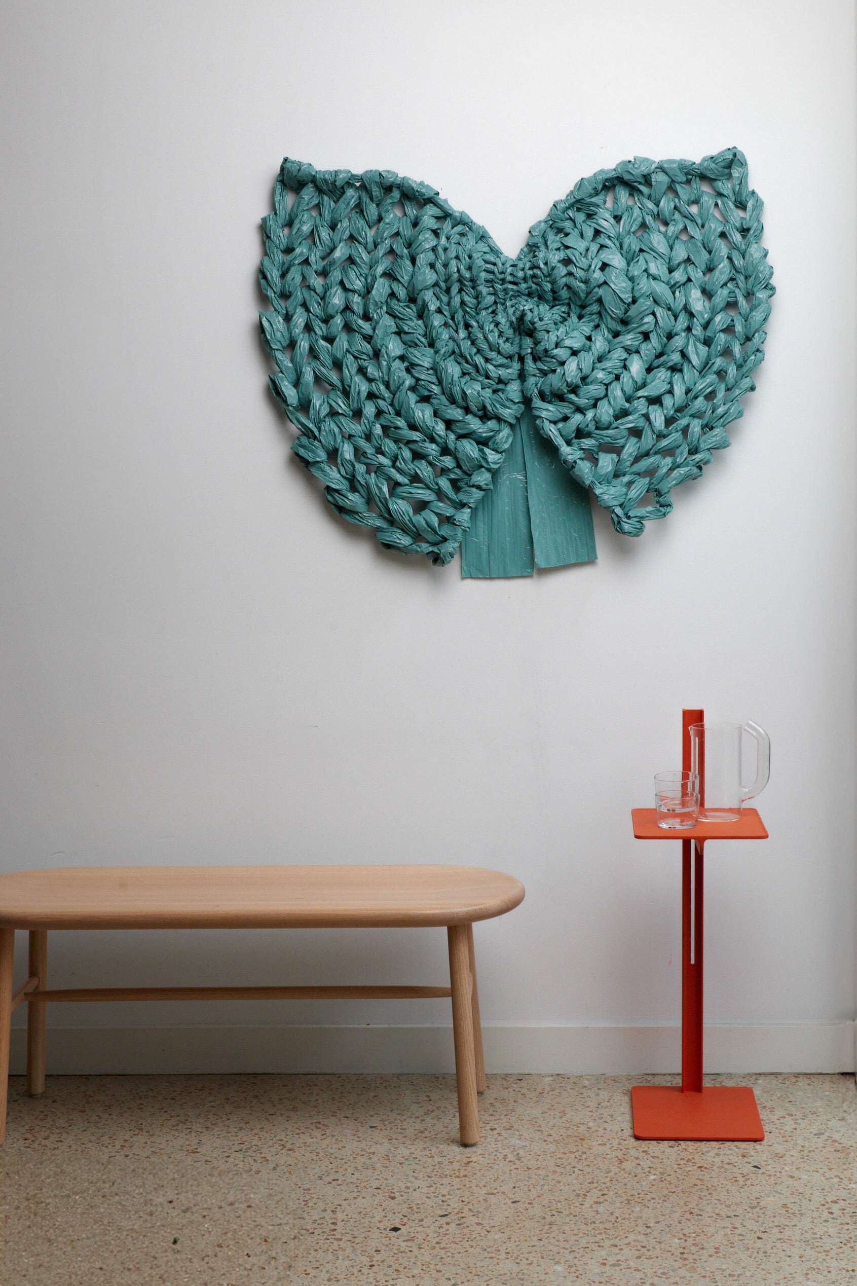 Teal knitted wall art piece on a white wall with a wooden bench and red stand in the foreground.