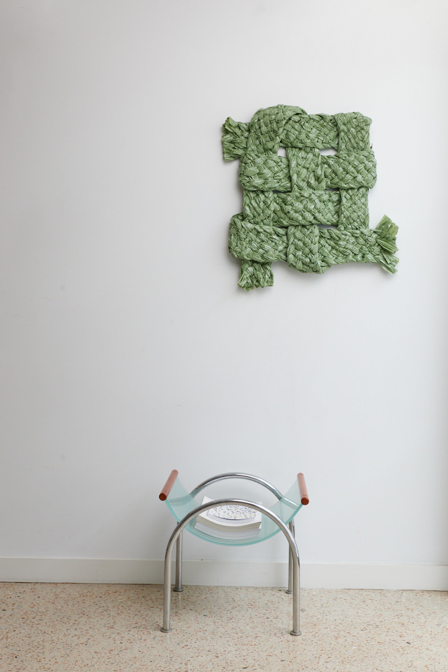 Green knitted wall hanging on a white wall with a chair in the foreground.